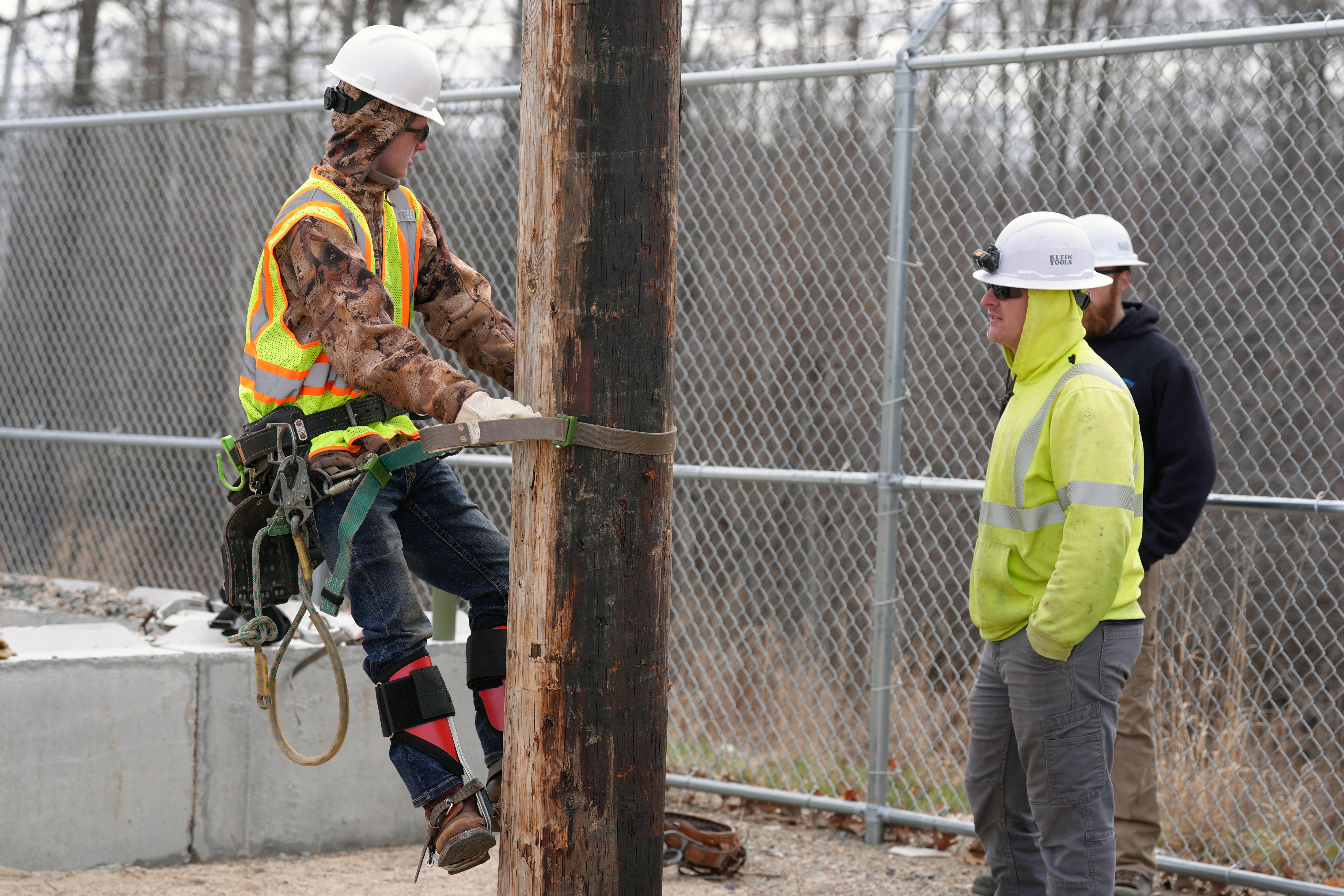A utility lineworker wearing a hard hat, high-visibility vest and climbing gear practices working on a wooden utility pole while two other workers observe from the ground near a fenced training area.