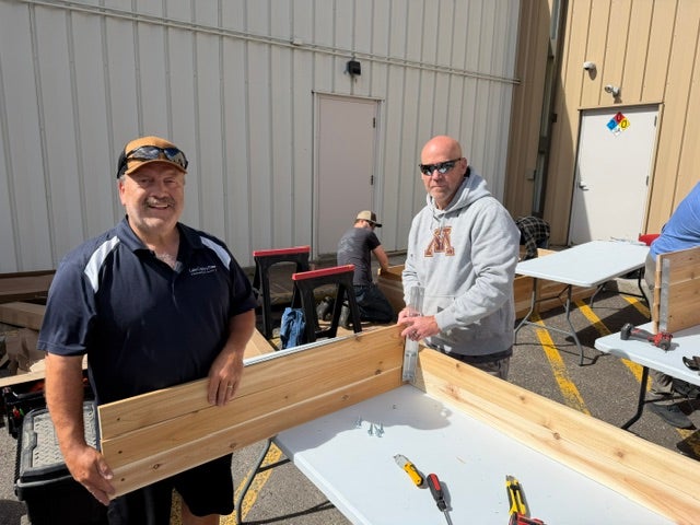 Two volunteers smile while assembling a wooden raised garden bed on a work table during a community service project.