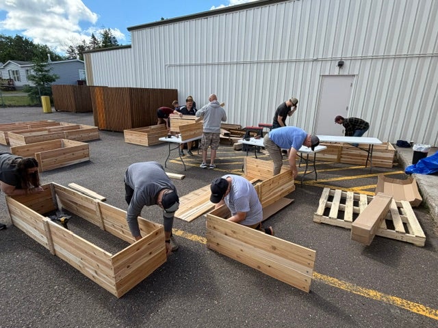Volunteers assemble wooden raised garden beds outdoors beside a large building, with lumber, pallets and tools spread throughout the work area.