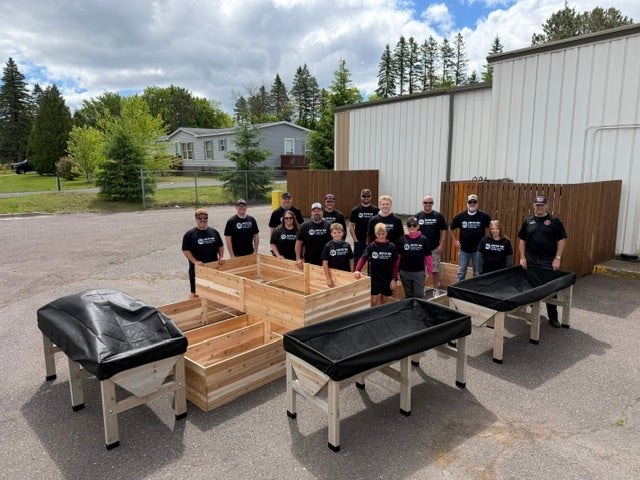 A group of volunteers poses behind newly constructed wooden raised garden beds and elevated planting tables during a community service project.
