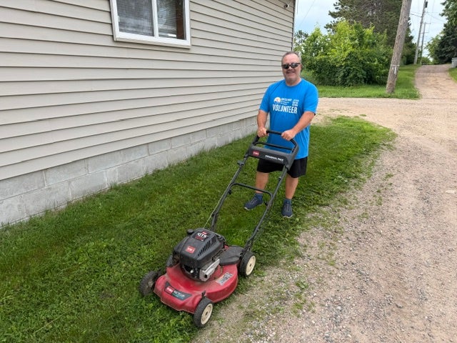 A volunteer in a blue United Way Day of Action shirt pushes a lawn mower while doing yard work alongside a home.