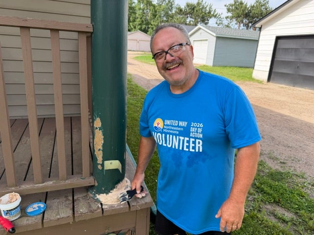 A volunteer in a blue United Way Day of Action shirt smiles while painting a green porch column at a home.