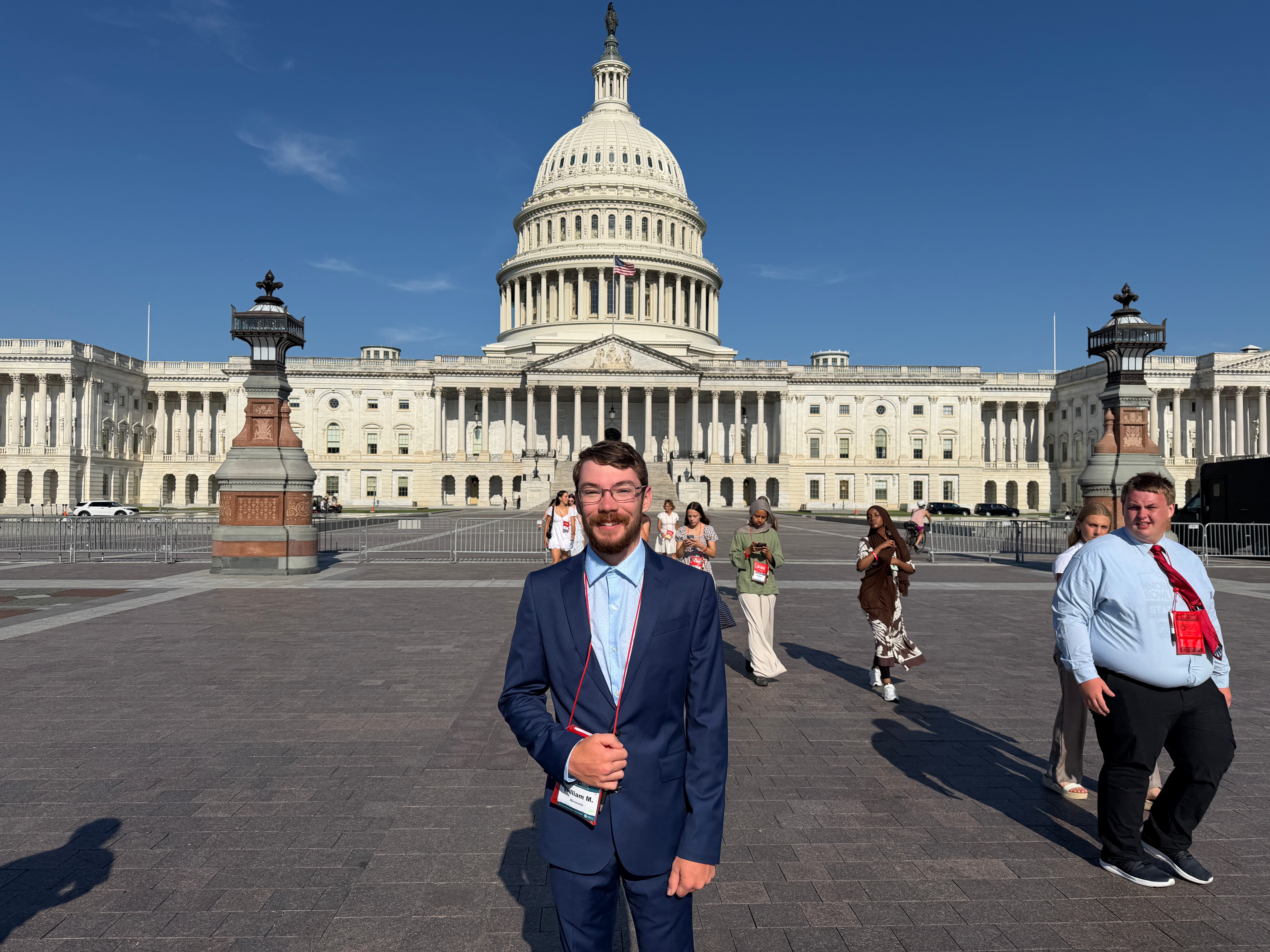 A young man in a navy suit smiles for a photo in front of the U.S. Capitol in Washington, D.C., as other visitors walk across the plaza behind him.