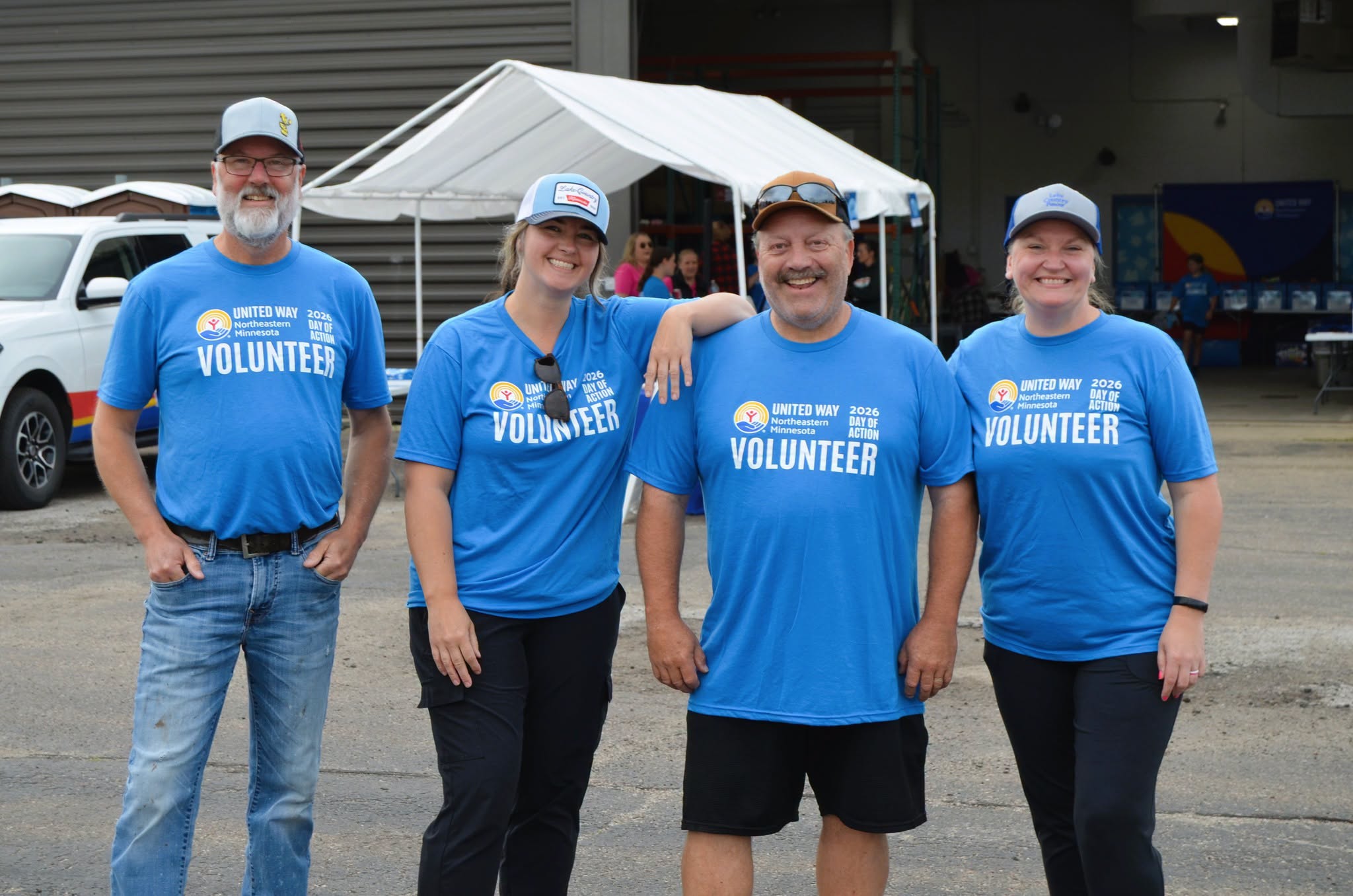 Four volunteers wearing blue United Way of Northeastern Minnesota Day of Action shirts smile together outside a volunteer work site.