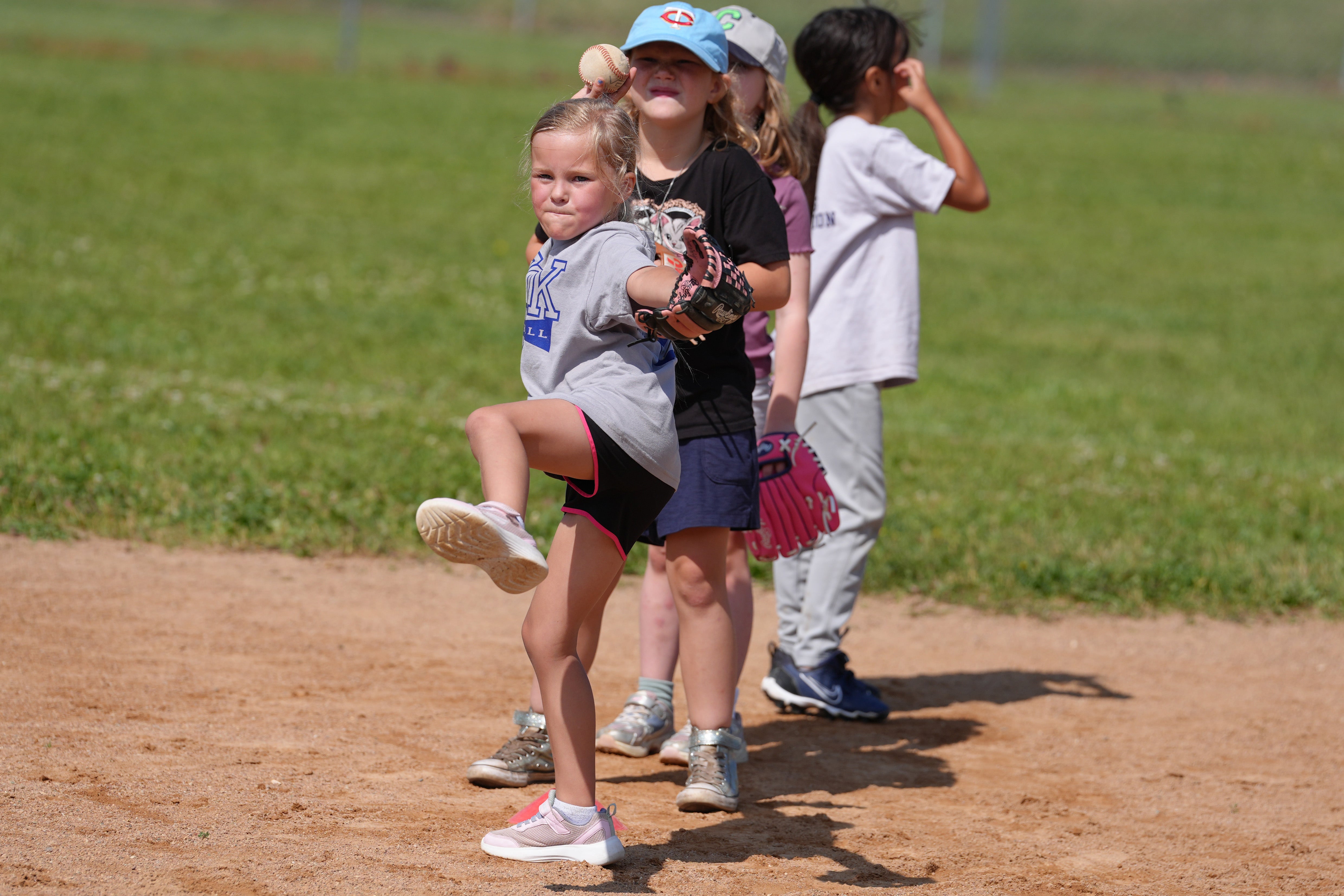 A young girl practices pitching on a baseball field, lifting one leg and preparing to throw as other children wait behind her.