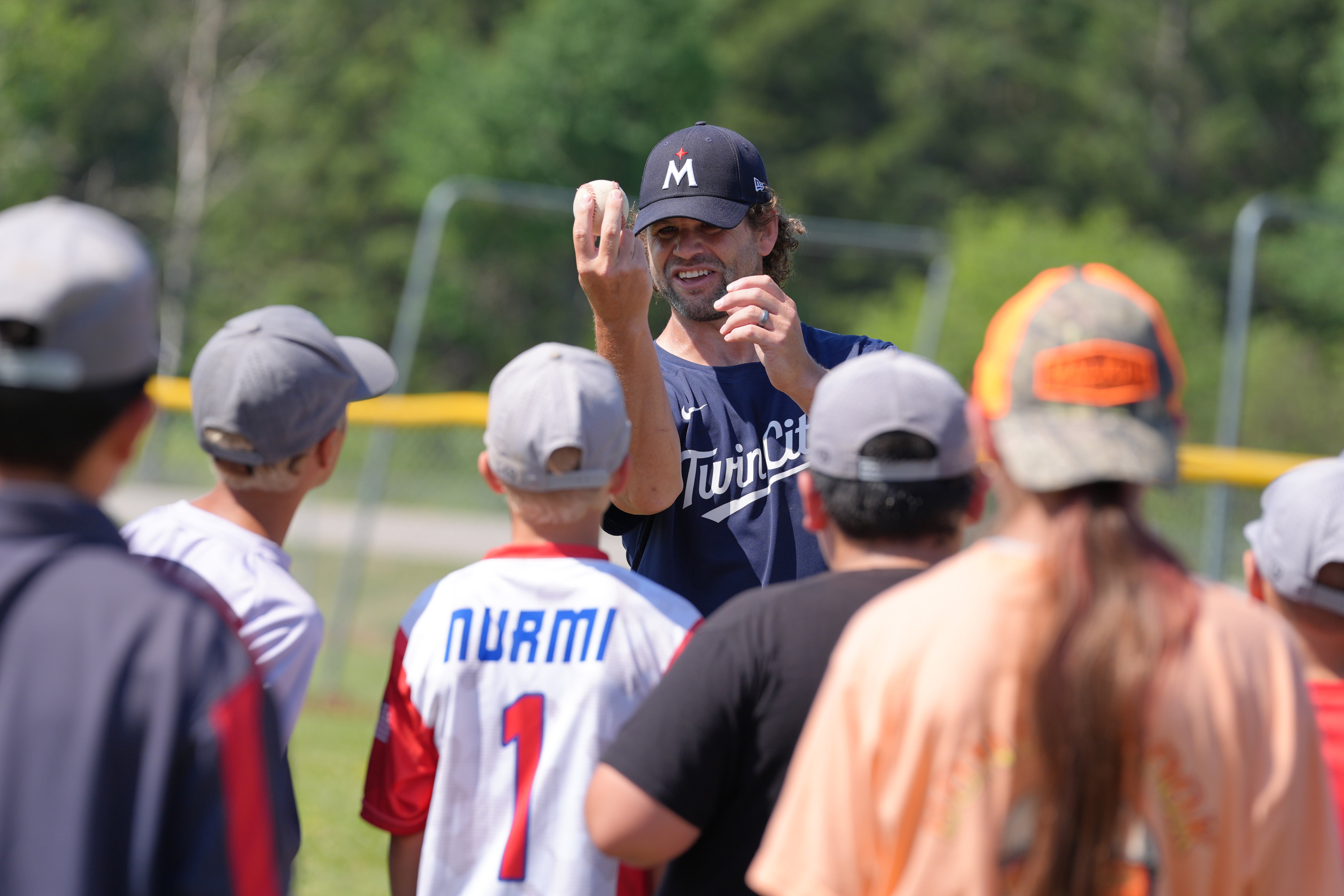 A Minnesota Twins clinic instructor holds up a baseball while demonstrating a technique to a group of young players gathered around him.