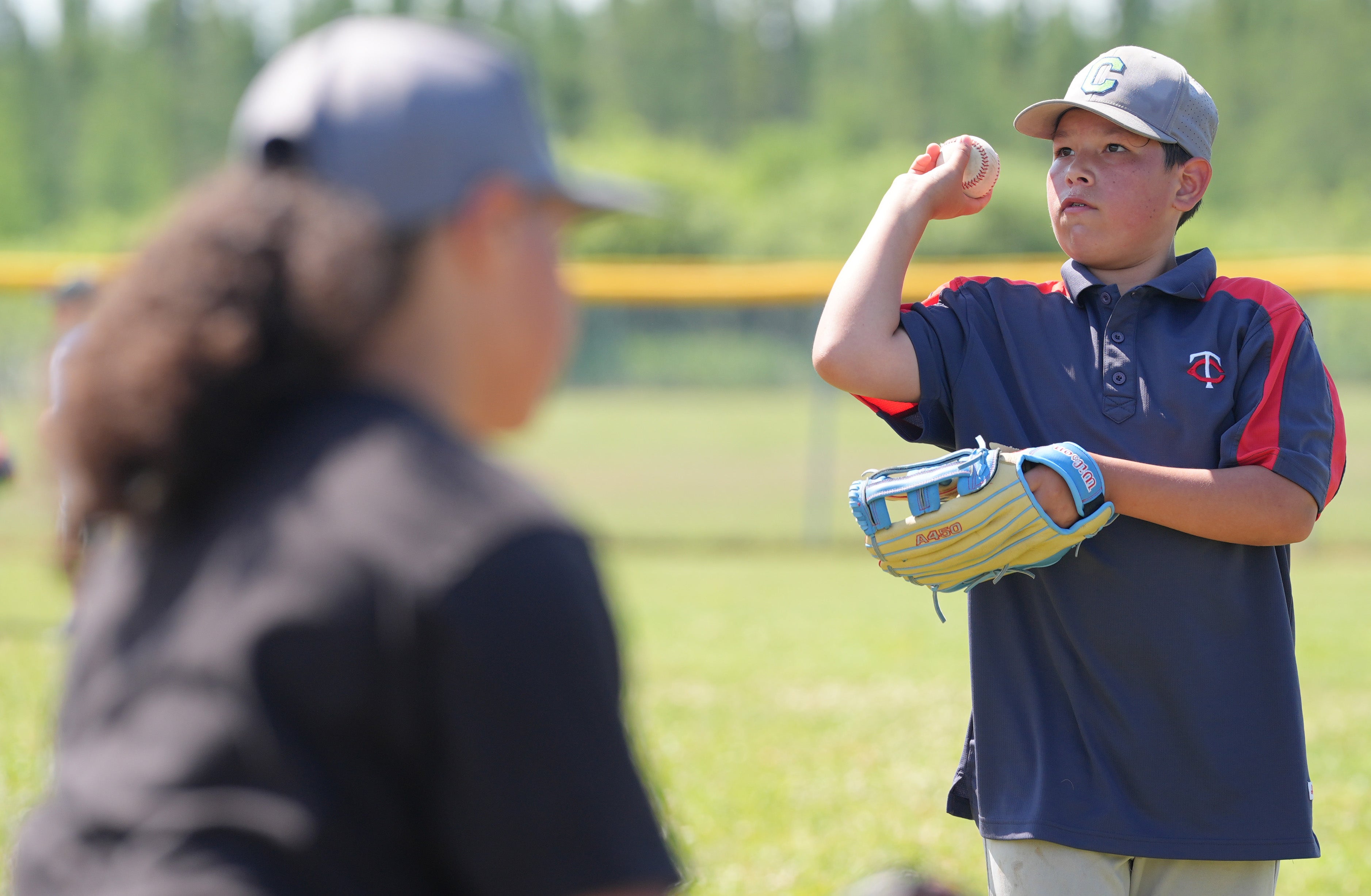 A young baseball player in a green jersey practices throwing a baseball while wearing a glove during an outdoor clinic.