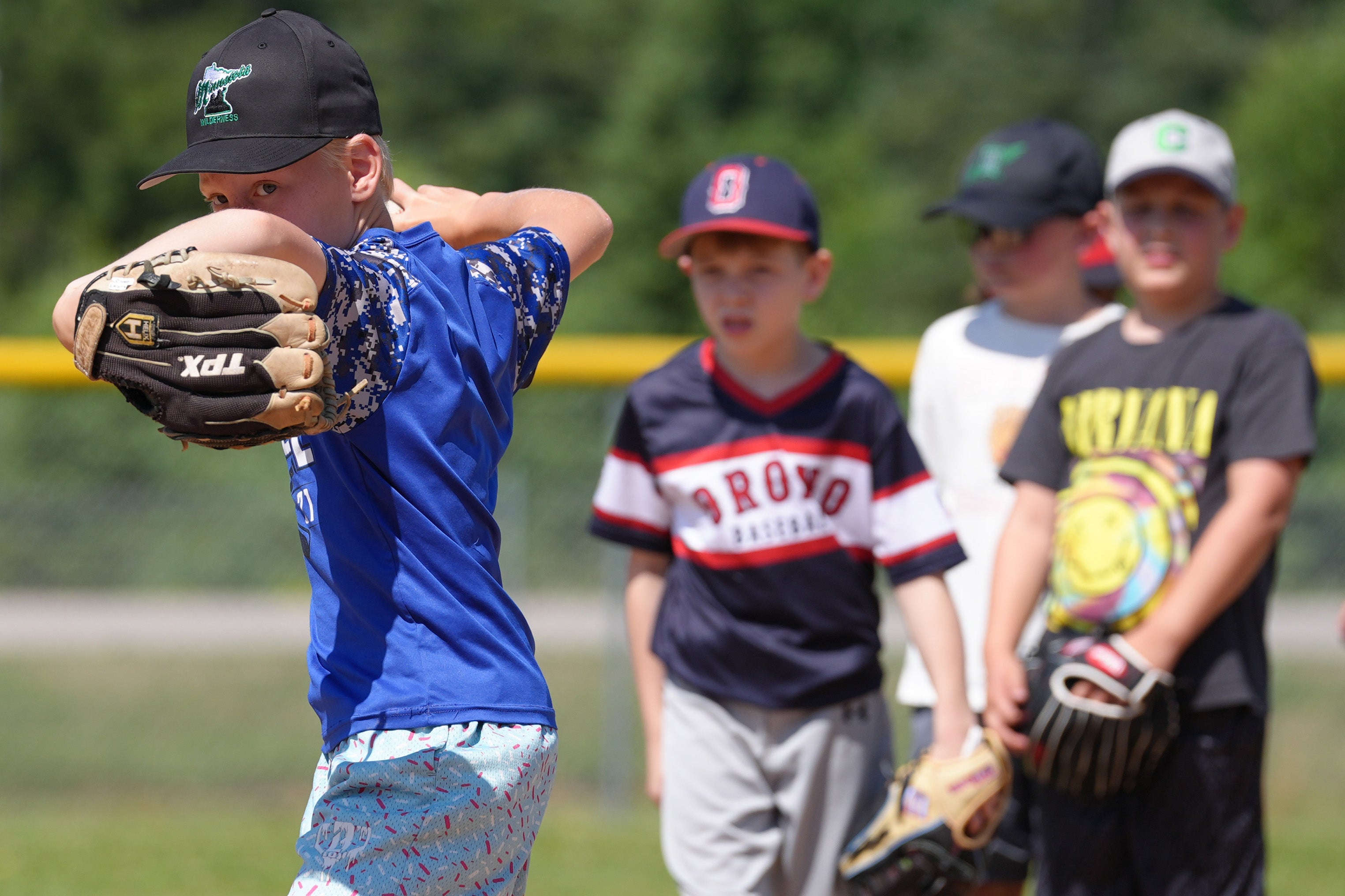 A young baseball player holds a glove in front of his face while practicing a pitching motion during an outdoor youth baseball clinic, with other children watching in the background.