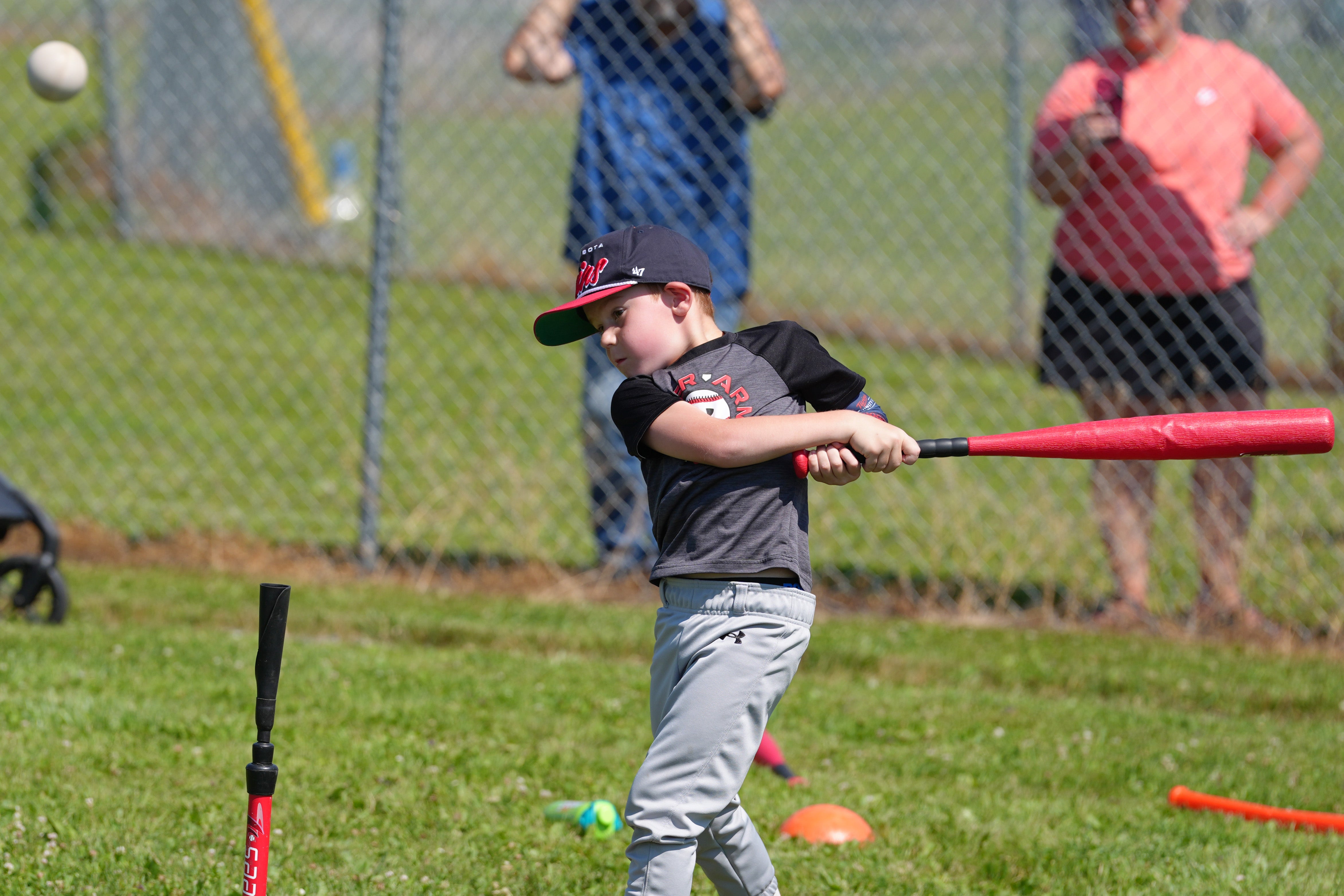 A young baseball player swings a red bat at an incoming ball during a hitting drill at an outdoor youth baseball clinic.