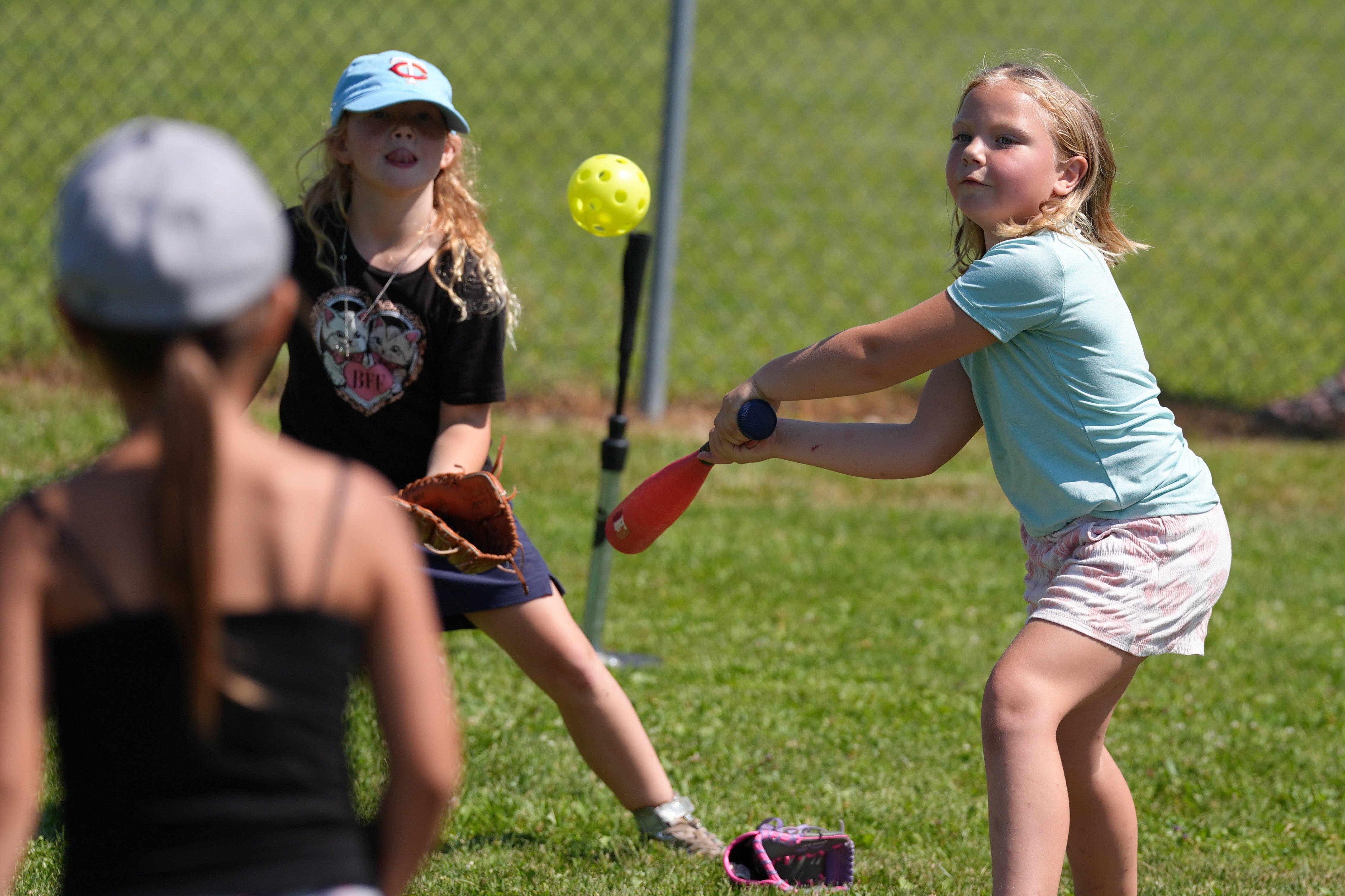 A young girl swings a red bat at a yellow practice ball during an outdoor youth baseball clinic while other children watch nearby.