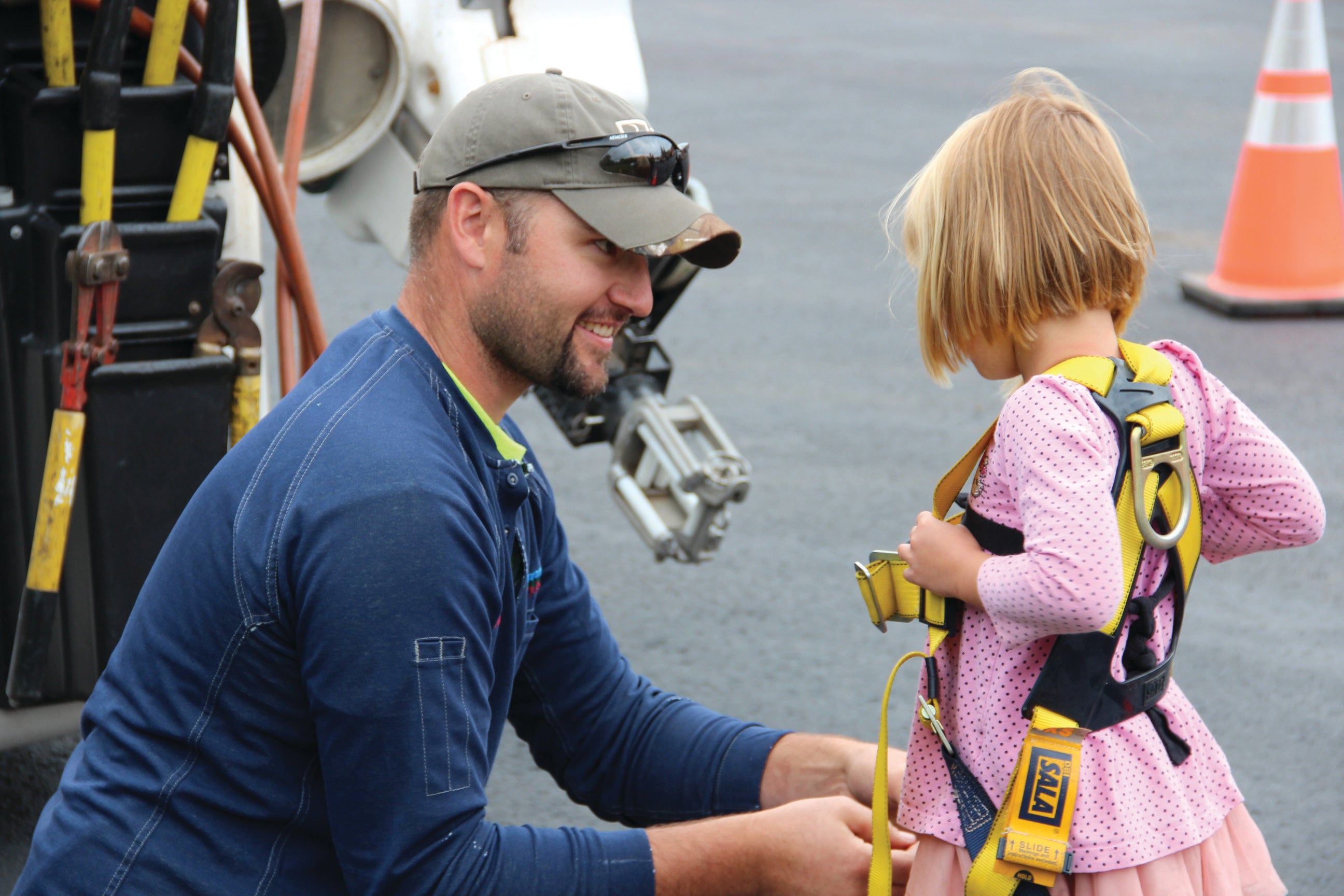 A utility worker kneels beside a young girl as she tries on a yellow safety harness near a utility truck. The smiling worker helps adjust the equipment while the child, dressed in pink, explores the safety gear during an outdoor demonstration.
