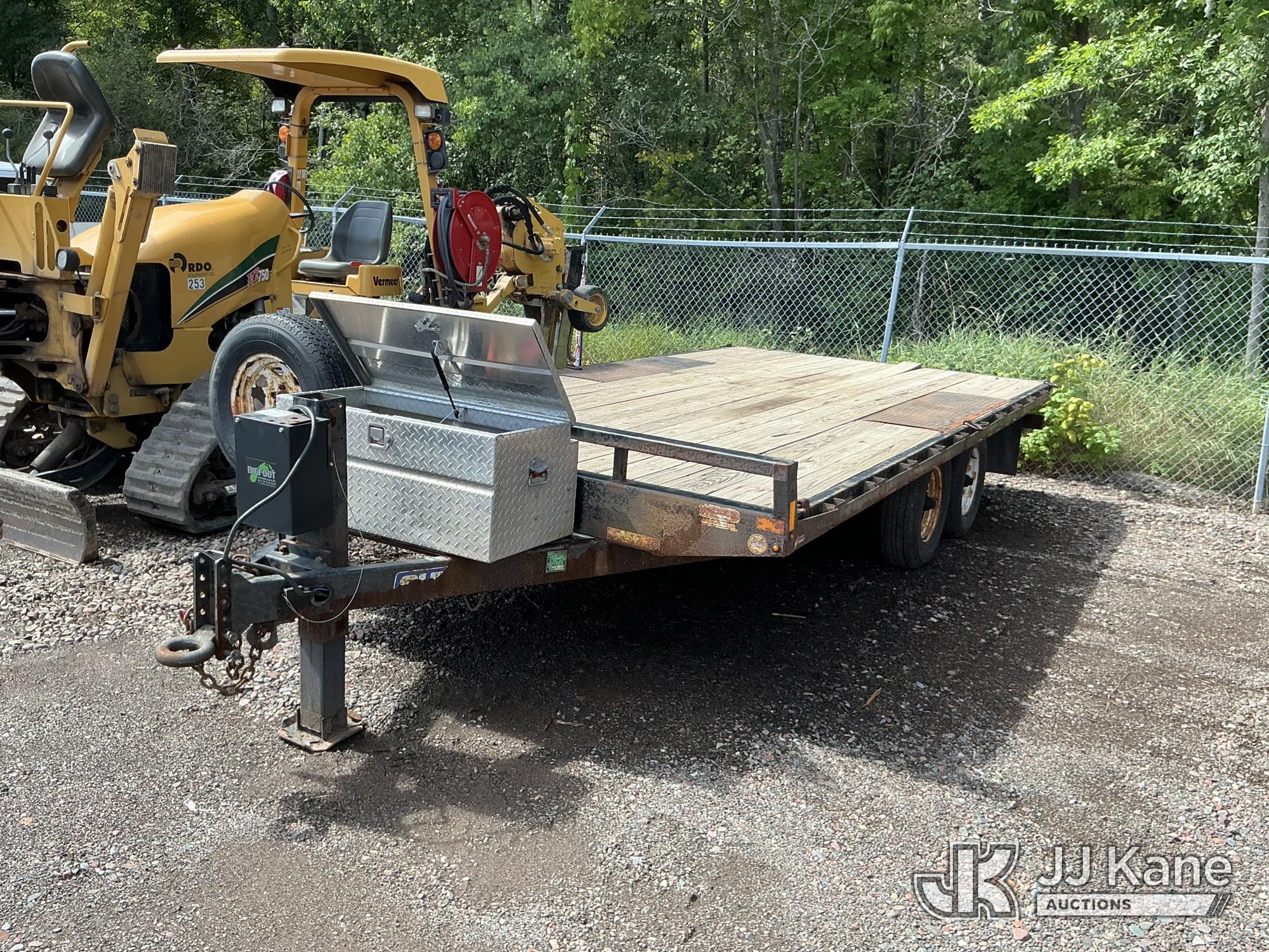 Tandem-axle Sure-Trac flatbed equipment trailer with a weathered wood deck, diamond-plate toolbox, hitch, safety chains and front jack, parked beside a chain-link fence.