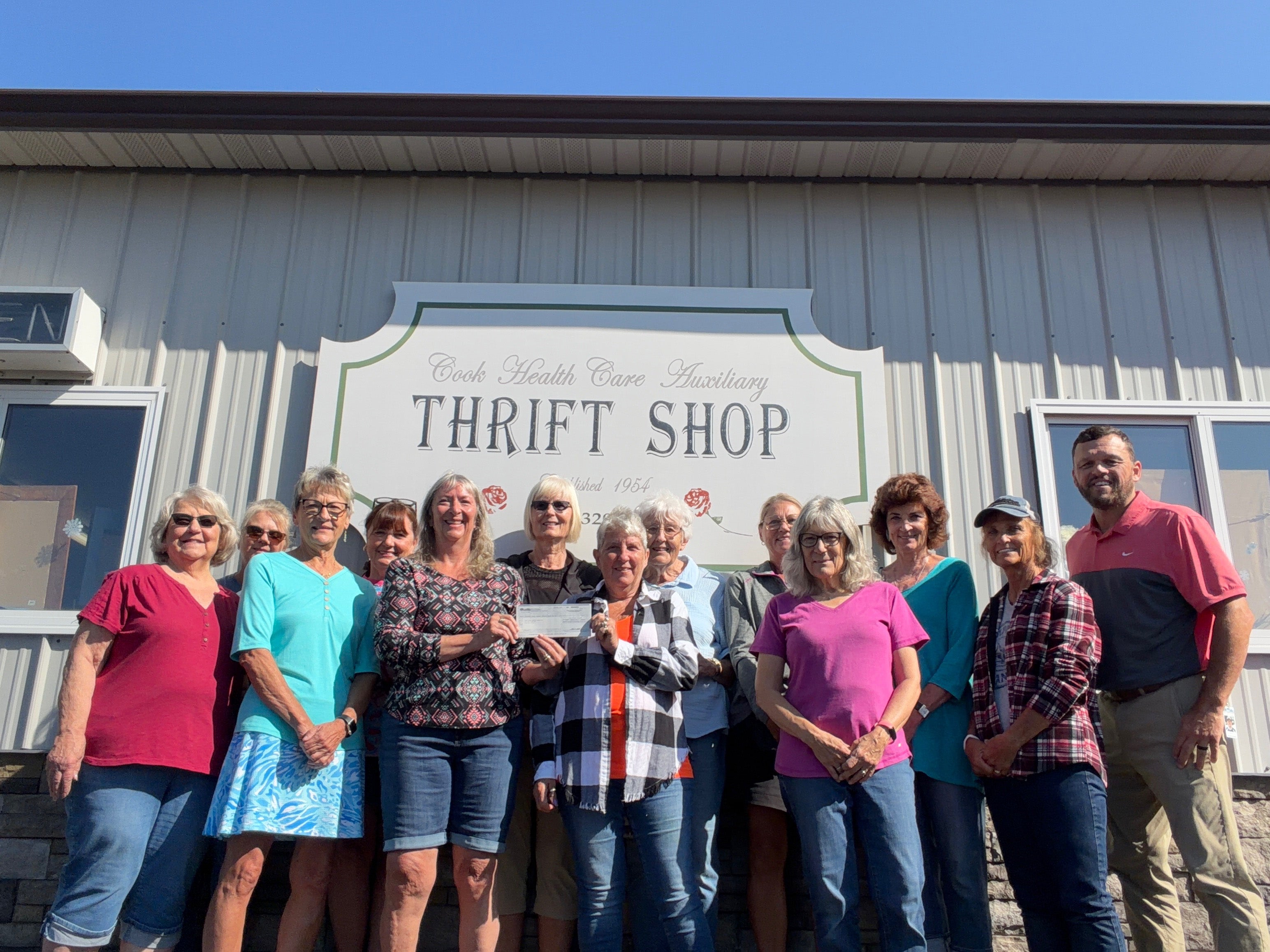 A group of volunteers and a Lake Country Power representative pose outside the Cook Health Care Auxiliary Thrift Shop while two women hold a donation check.