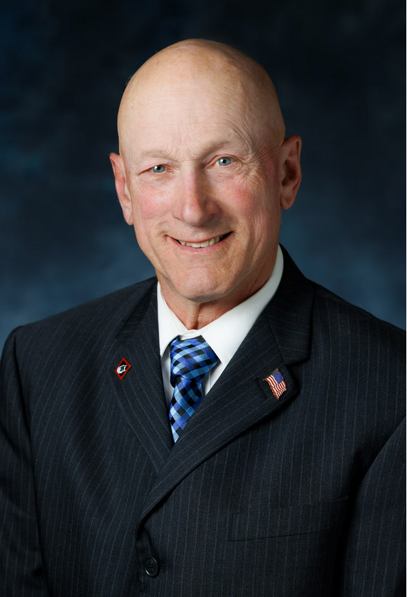Portrait of a smiling bald man wearing a dark suit, white shirt and blue patterned tie, with an American flag pin on his lapel against a dark blue background.