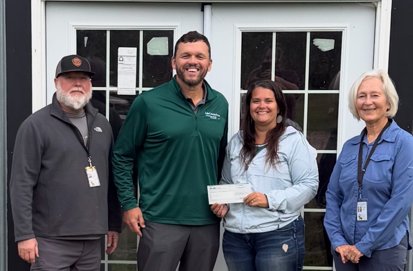 Four people stand together outside a building, smiling as a woman in the center holds a check presented to her.