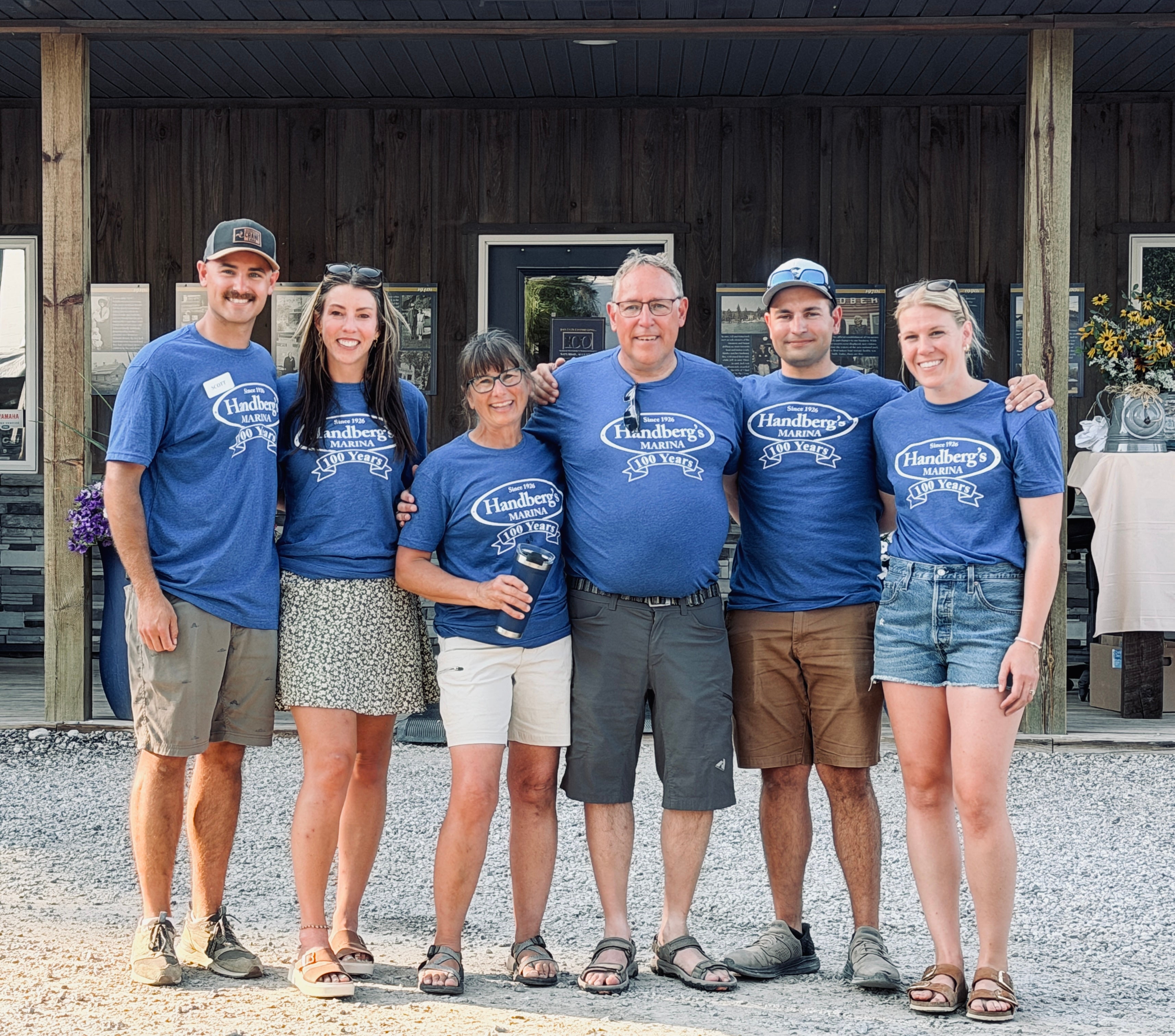 Six people wearing matching blue Handberg’s Marina 100 Years shirts pose together outside the marina.