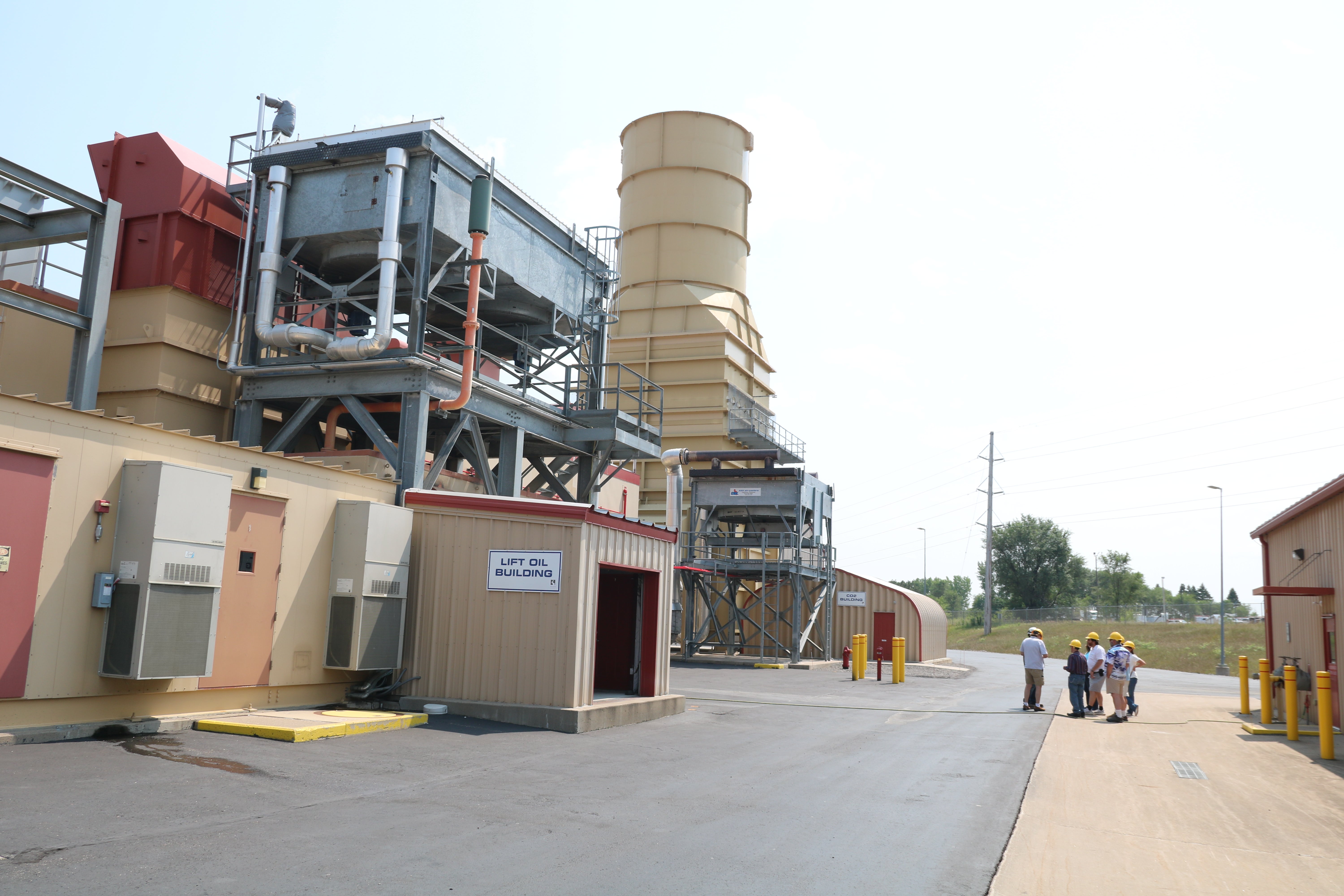 A group wearing hard hats tours an industrial facility featuring large processing equipment, piping and a tall exhaust stack.
