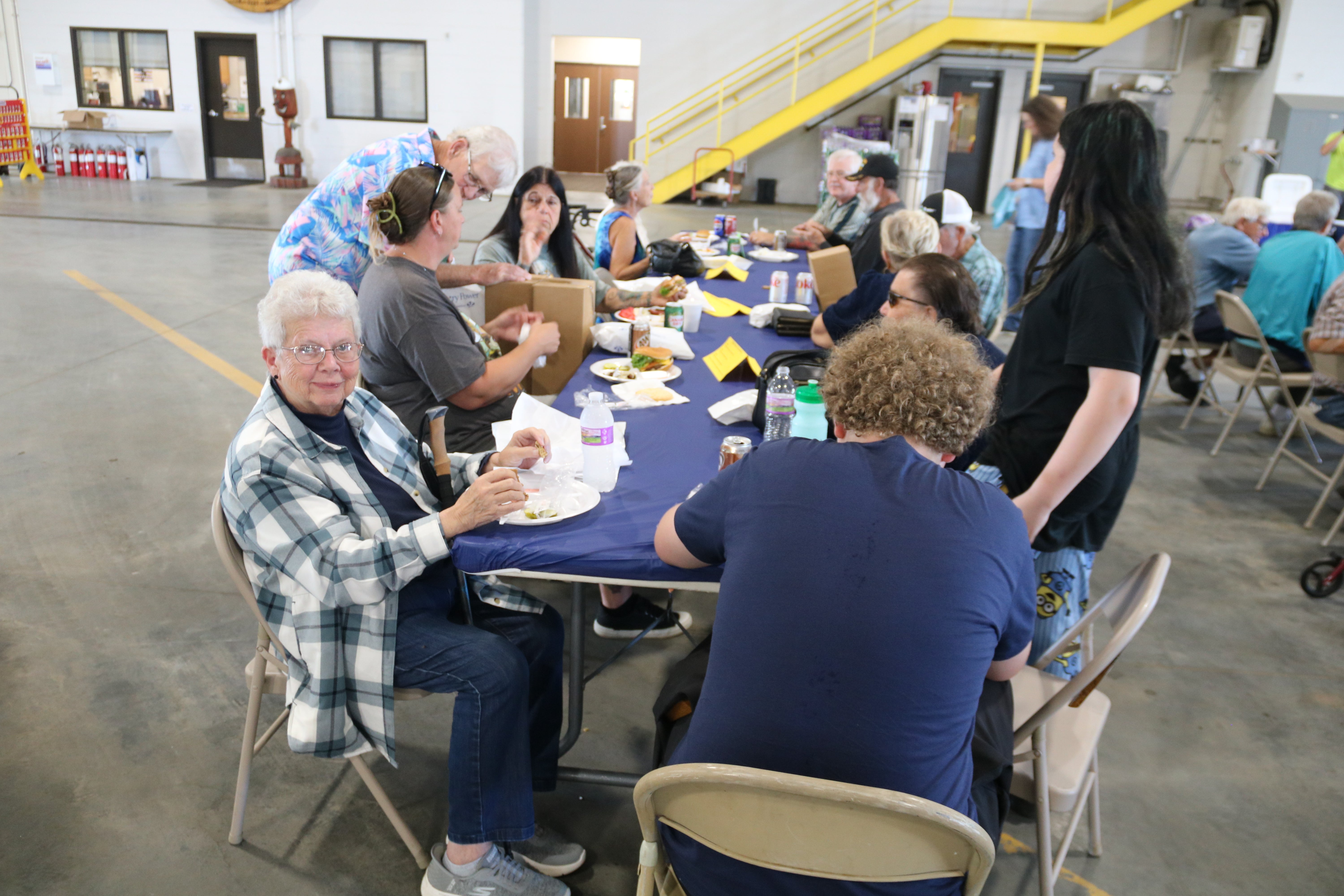 Community members gather around a long table for a meal and conversation inside a large utility facility, with several people seated and others standing nearby.