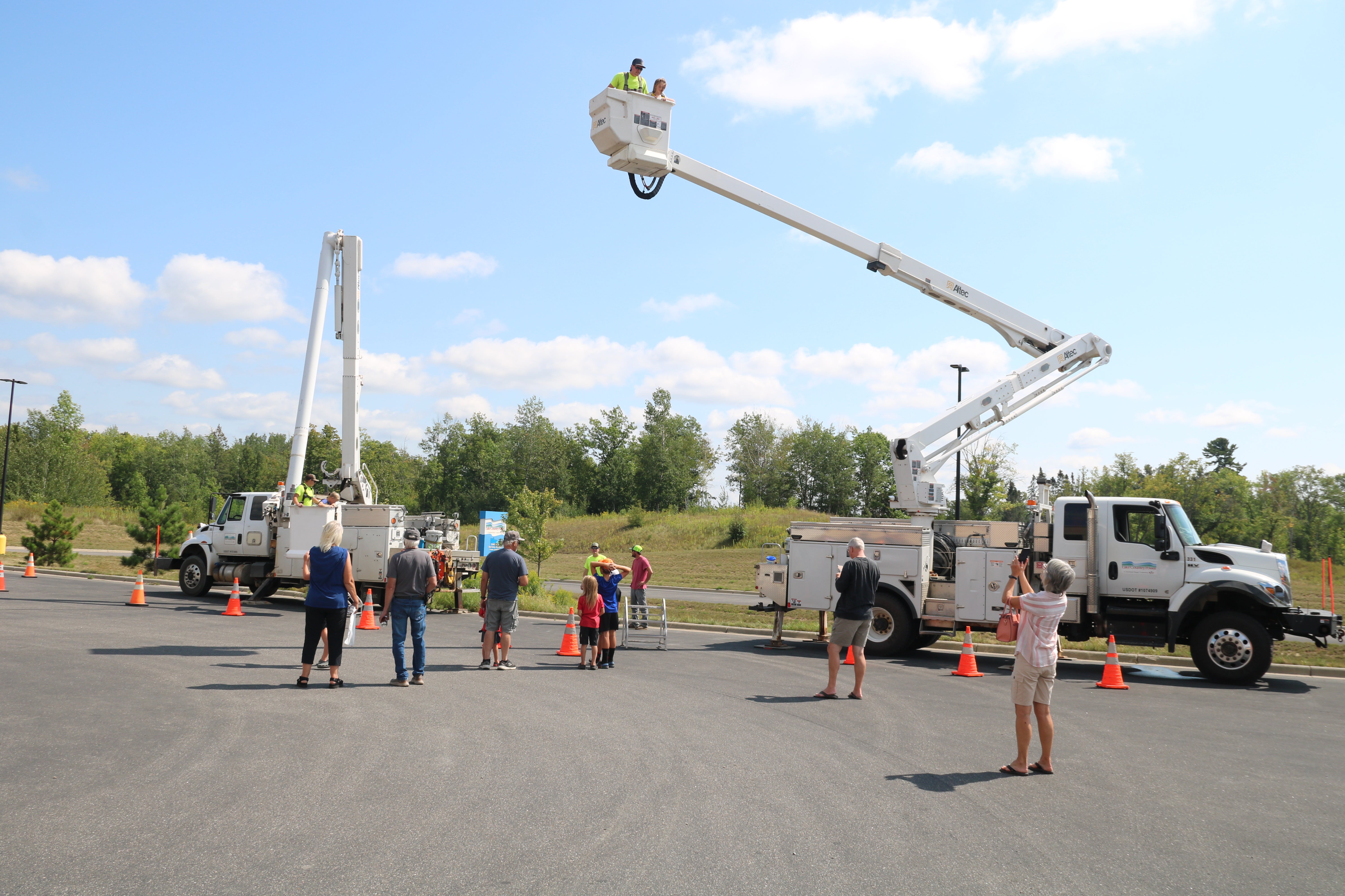 Lake Country Power bucket trucks on display during an outdoor community event, with visitors watching as workers and a child ride high above the ground in an extended bucket lift.