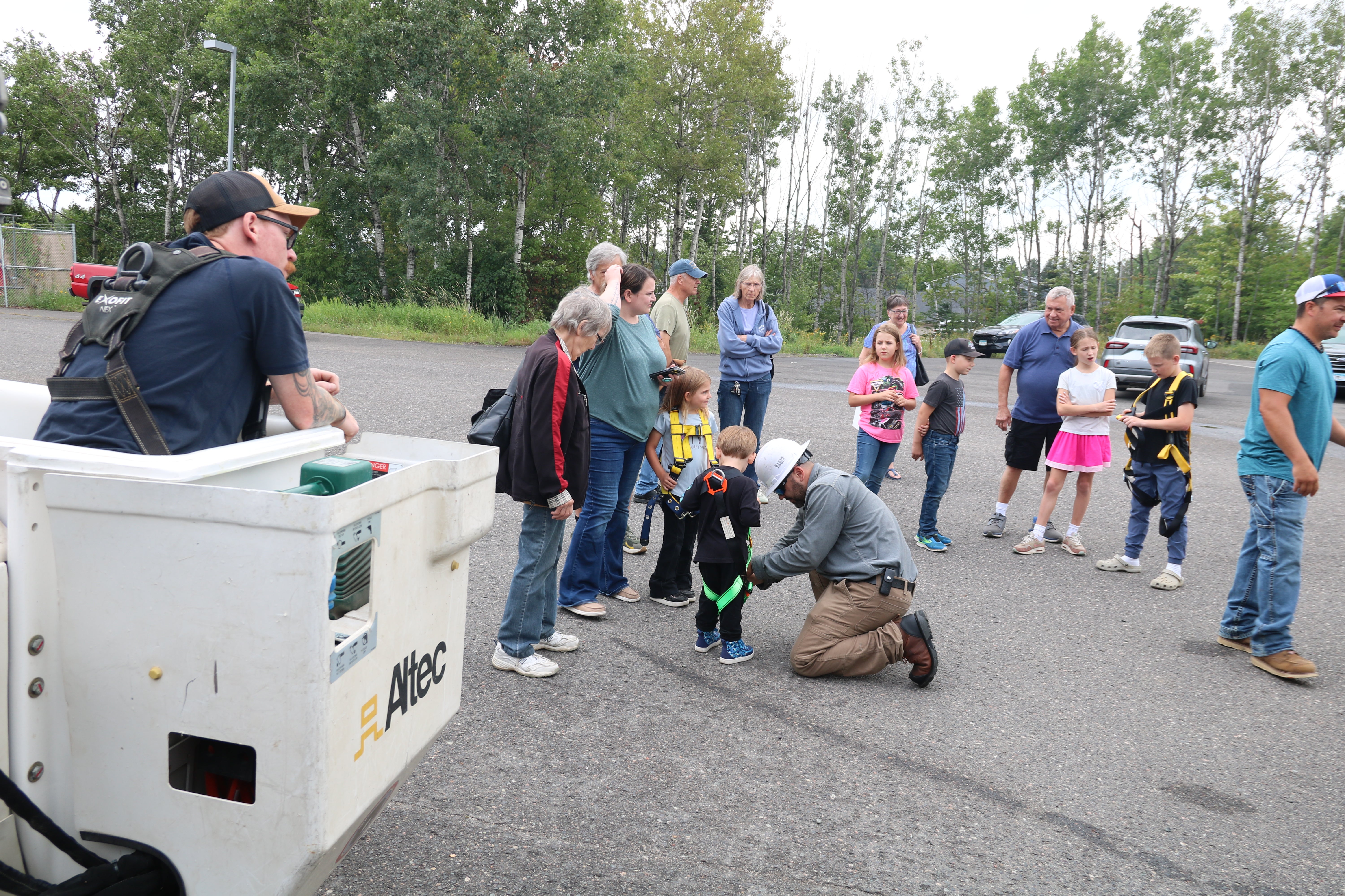Families gather around a utility bucket truck during a community event as a worker helps a young child put on a safety harness while others wait nearby.