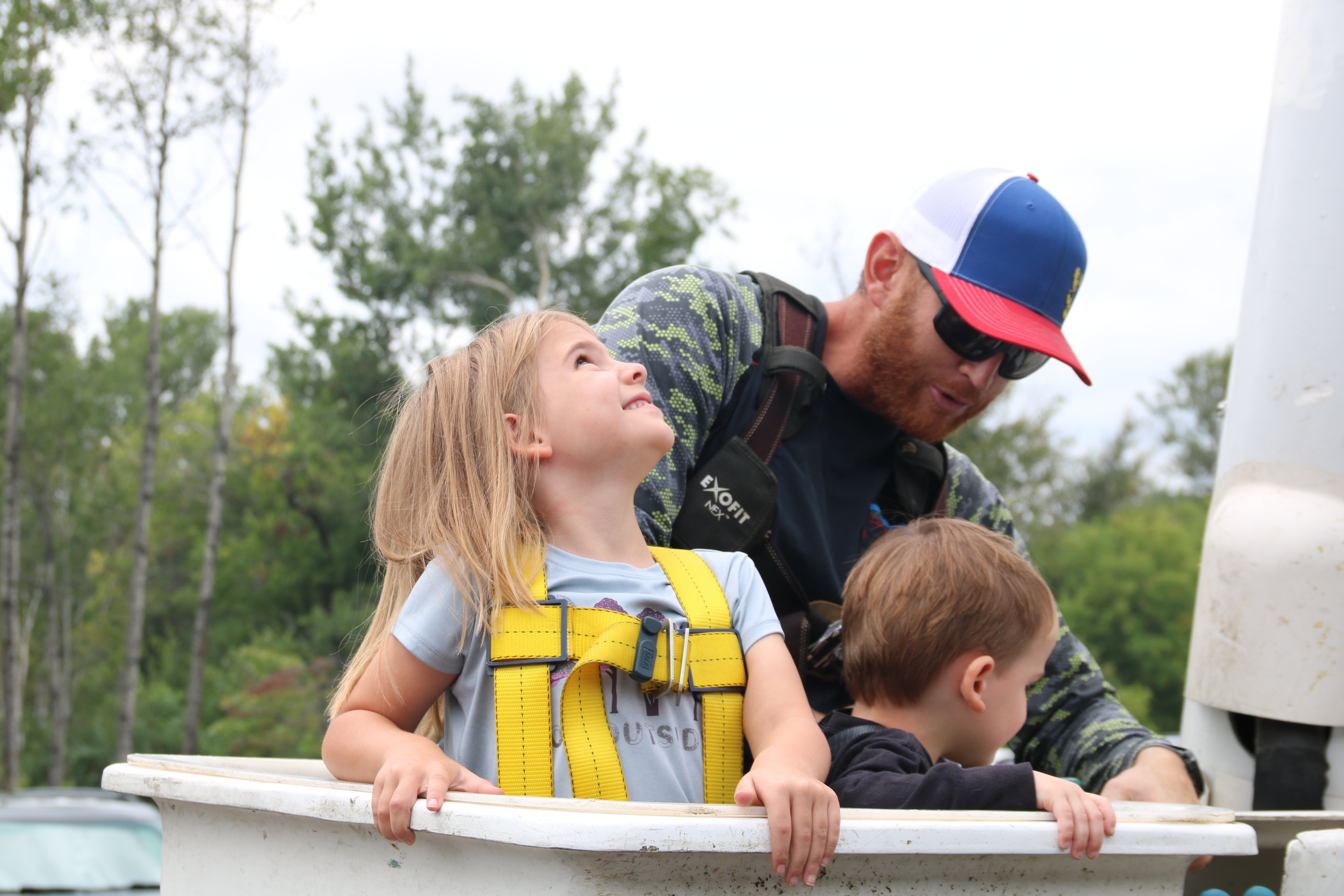Two young children wearing safety harnesses stand in a utility bucket while a worker helps prepare them for a bucket truck ride at an outdoor community event.