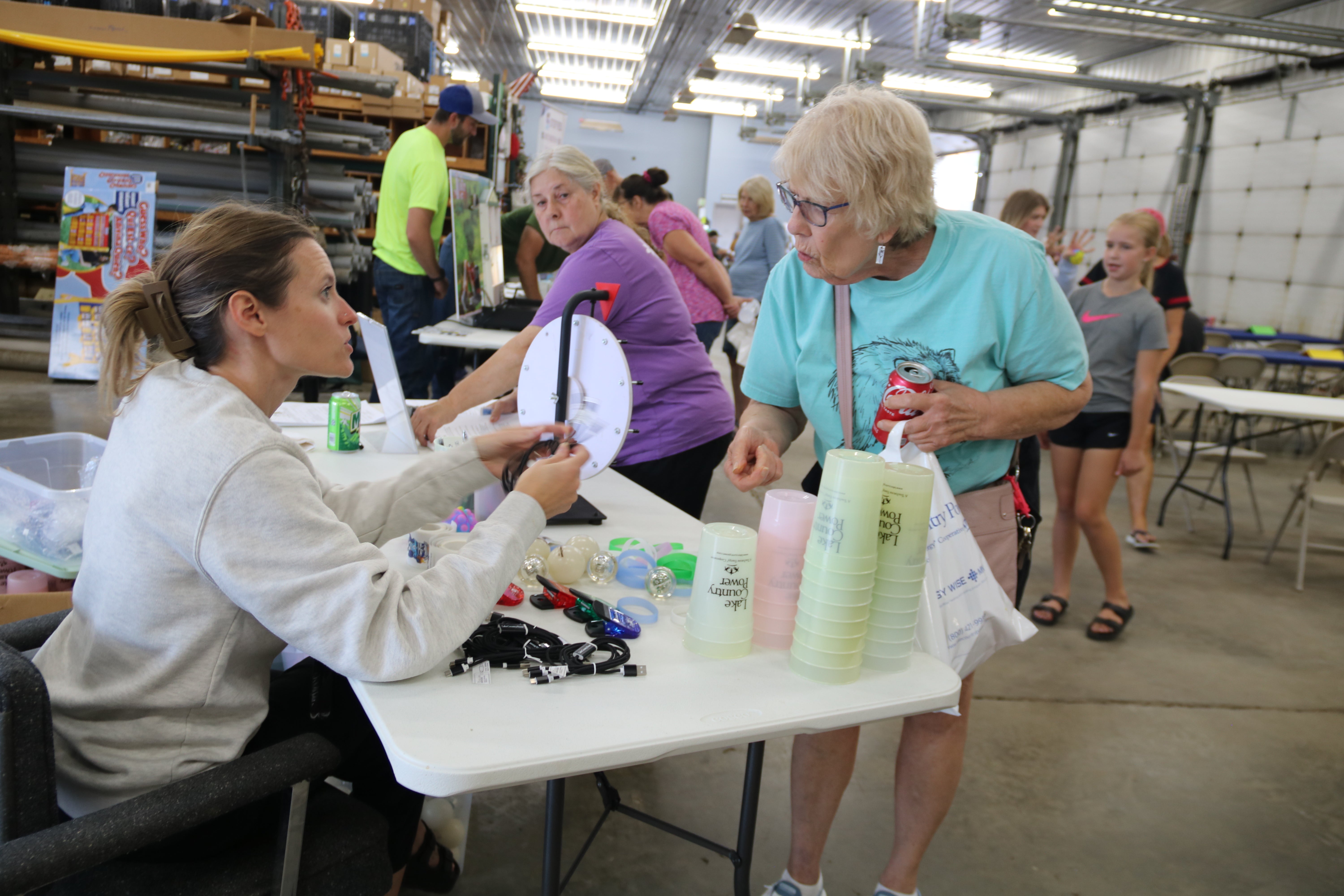 A Lake Country Power employee assists an attendee at a giveaway table during a community event, with cups, small promotional items and charging cables displayed on the table.