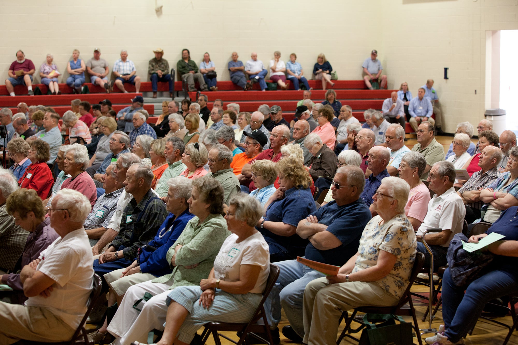 A large crowd of community members sits in folding chairs and bleachers during a public meeting or gathering in a gymnasium.